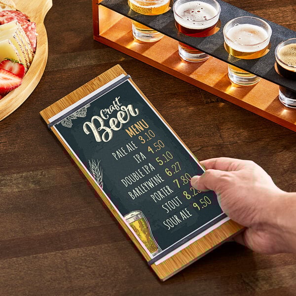 A hand holding a menu in a natural wood color menu holder on a table with a glass of beer.