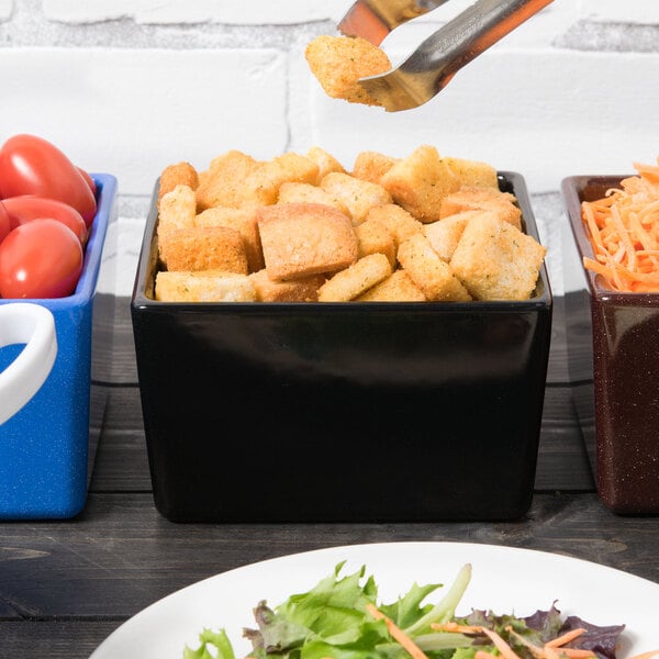 A black Tablecraft bowl filled with croutons on a table with a salad plate.