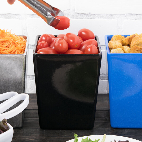 A person uses a knife to cut a tomato at a table into a black square container of cherry tomatoes.