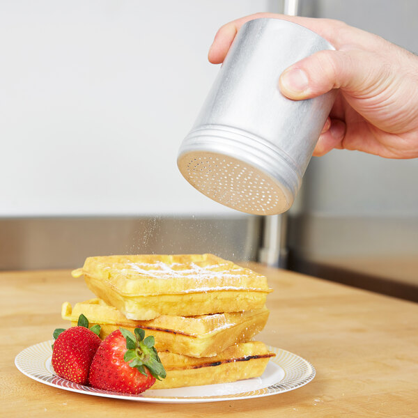 A hand pouring powdered sugar from a silver Vollrath shaker onto a stack of waffles.