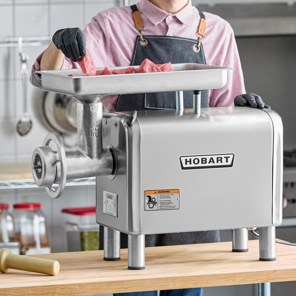 A commercial Hobart meat grinder and chopper with a stainless steel body, shown in use with raw meat pieces in a kitchen setting.