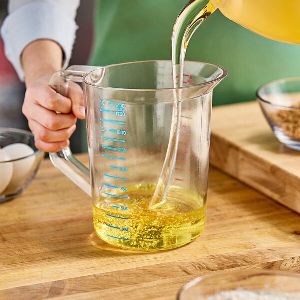 A person pouring yellow liquid into a Rubbermaid clear polycarbonate measuring cup on a kitchen counter.