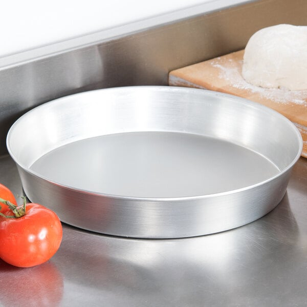 An American Metalcraft tin-plated steel deep dish pizza pan with a tomato next to dough on a cutting board.