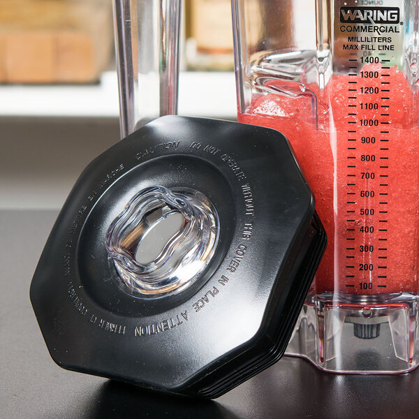 A black Waring lid on a clear plastic container with a measuring cup