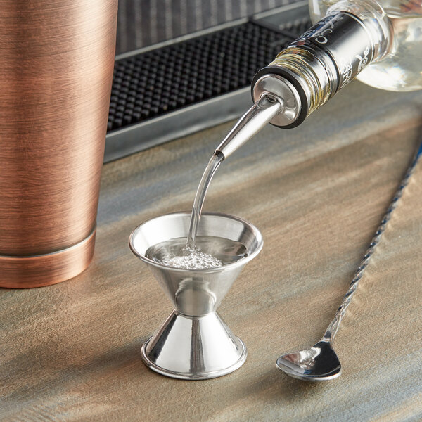 An American Metalcraft stainless steel jigger on a table with a cocktail shaker and a bottle.