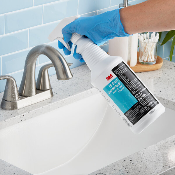 A person in blue gloves using 3M TB Quat Disinfectant spray to clean a white sink.