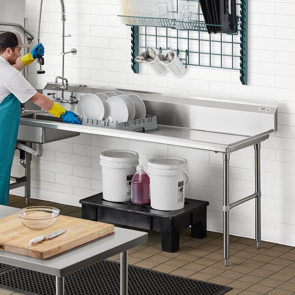 A man in overalls and an apron washing dishes in a commercial kitchen with a Regency dirty dish table.