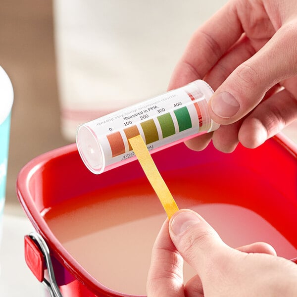 A vial of Noble QT-10 quaternary test strips being used to test the concentration of a liquid in a red container.