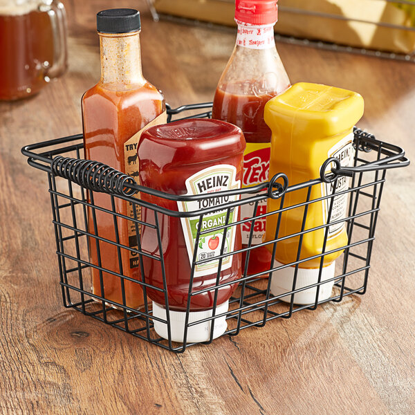 An American Metalcraft black metal tabletop market basket filled with bottles of ketchup, mustard, and mustard on a table.