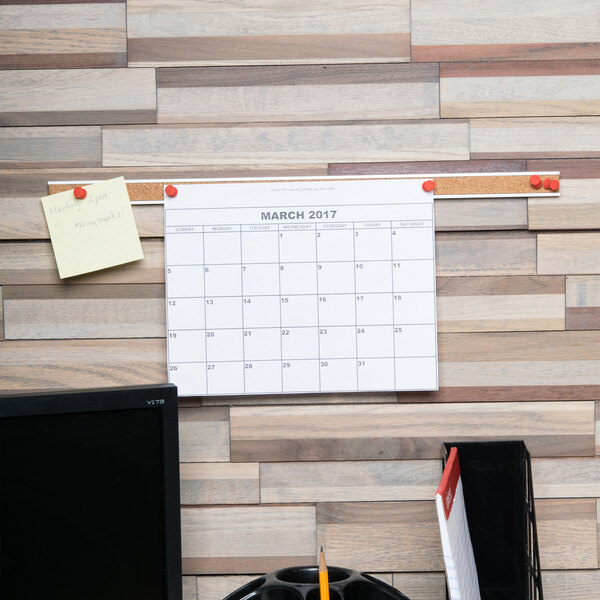 A brown cork bulletin bar on a wall with a calendar.