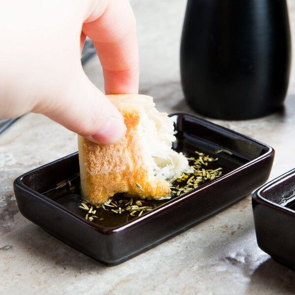A person holding a piece of bread in a small black Tuxton sauce dish with liquid.