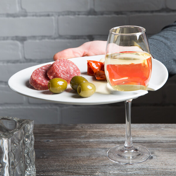 A person holding a Tuxton Bright White China appetizer plate with a glass of wine and vegetables.