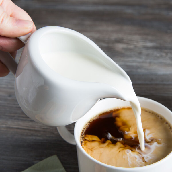 A person pouring milk into a cup of coffee using a Tuxton Bright White China creamer.