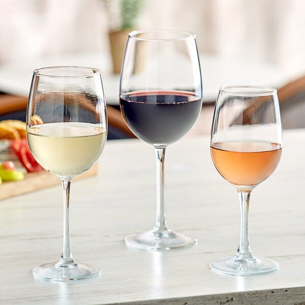 A clear 16 oz. wine glass filled with white wine, shown alongside glasses of red and rosé wine.