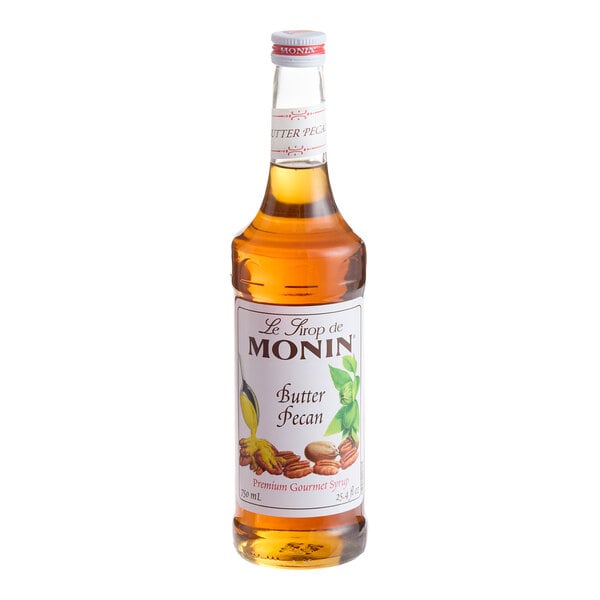 A clear glass bottle filled with amber-colored liquid, labeled as Monin Butter Pecan syrup with images of pecans and leaves on the front.