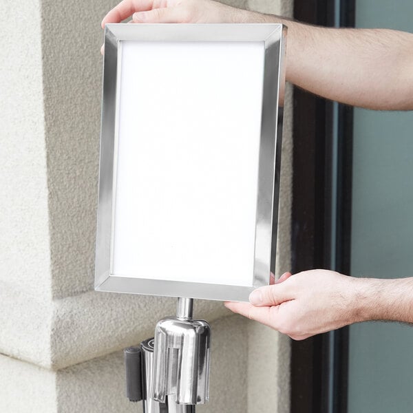 A person's arm holding a white sign in a silver Lancaster Table & Seating stanchion sign frame.