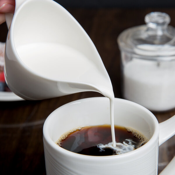 A person pouring white liquid from a white Royal Rideau porcelain creamer into a cup of coffee.