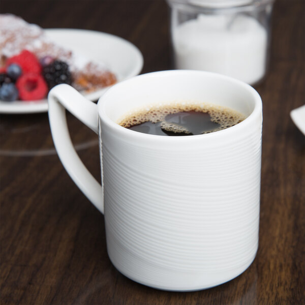 A white Reserve by Libbey Royal Rideau porcelain mug filled with brown liquid on a saucer next to a plate of food.