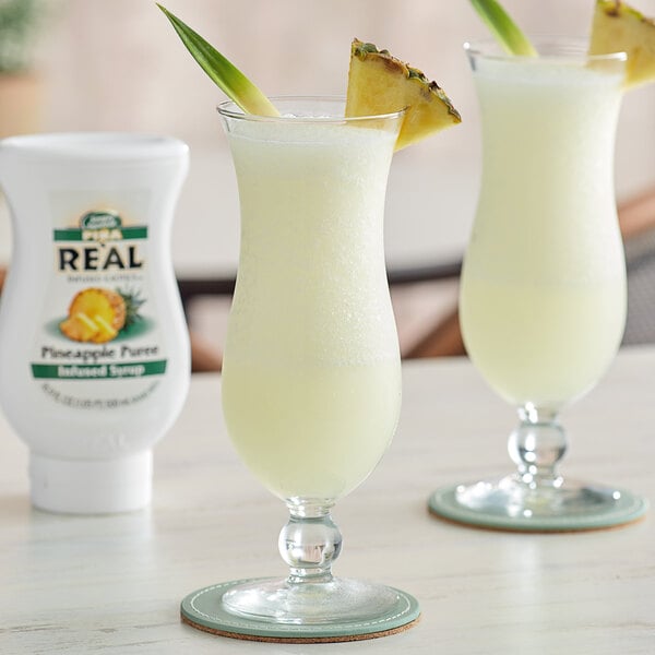 A close up of two glasses of yellow liquid with pineapple slices on top next to a white bottle of Real Pineapple Puree Infused Syrup.