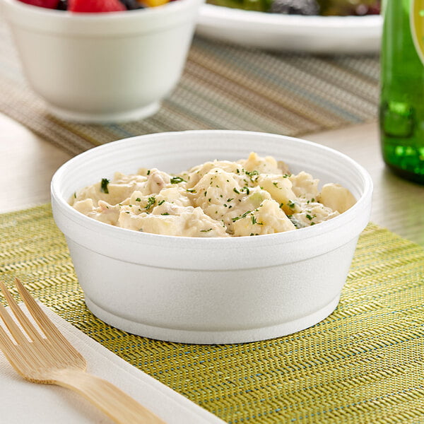 A Dart white foam bowl filled with food on a table with a napkin and wooden fork.
