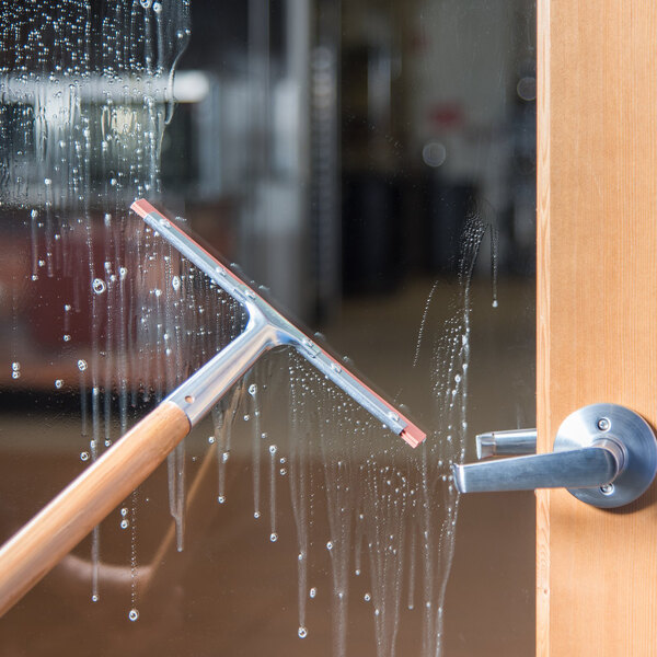 A window squeegee with a wooden handle and double rubber blade being used to clean a glass door.