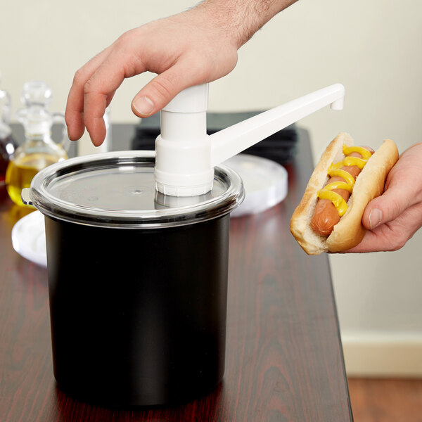 A hand holding a hot dog with mustard over a black plastic container with a white spout and cap.