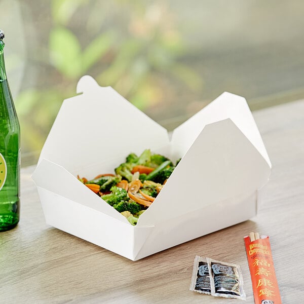 A white microwavable folded paper take-out container filled with food, shown on a table with condiments and chopsticks nearby.