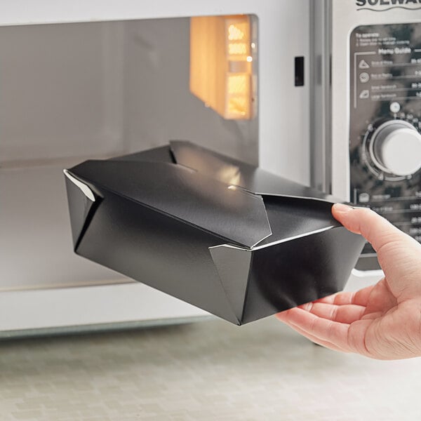 A hand holding a black Choice take-out box with a fold over lid in front of a microwave.