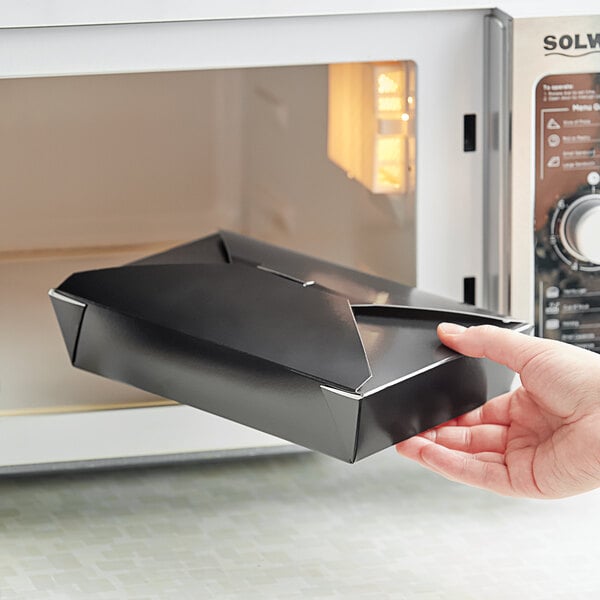 A hand holding a black Choice folded paper take-out box with a black lid coming out of a microwave.