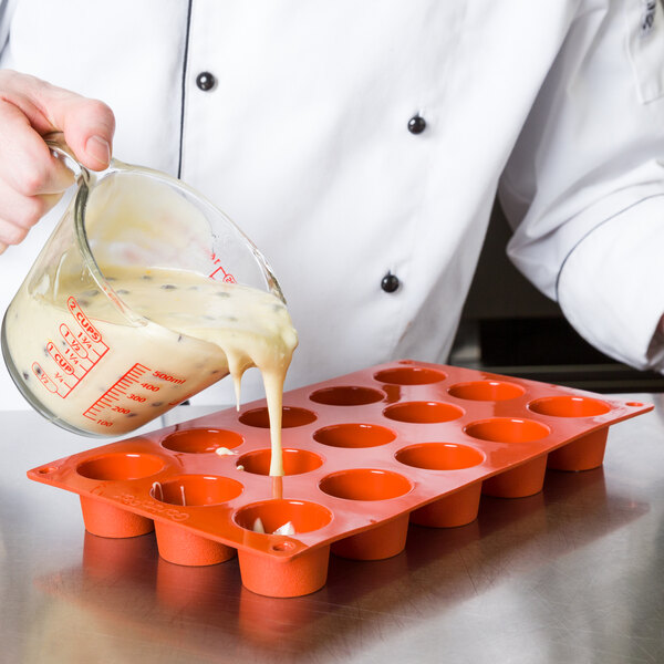 A chef using a Matfer Bourgeat orange silicone mini muffin mold to pour white liquid into cups.