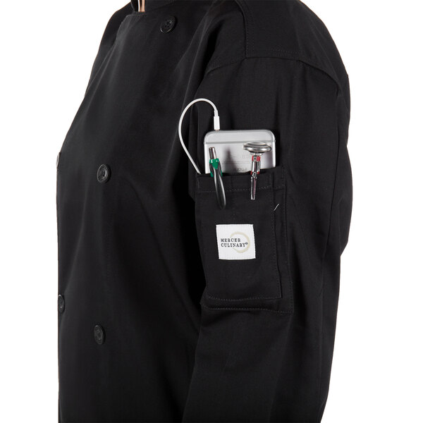 A person wearing a black Mercer Culinary chef jacket with a pocket full of tools on a professional kitchen counter.