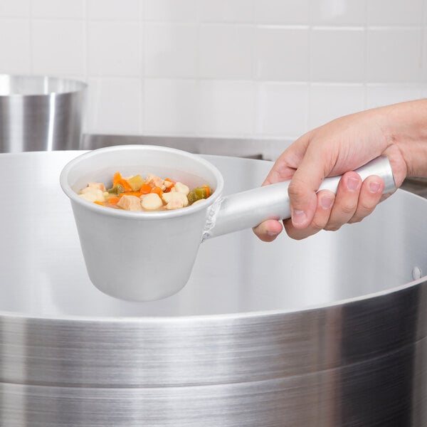 A person holding a Vollrath aluminum dipper over a pot of soup.