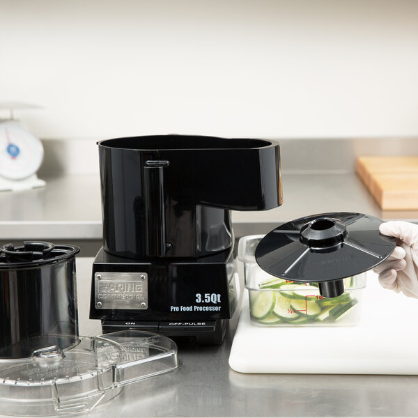A hand using a Waring continuous feed slinger to prepare vegetables in a professional kitchen.