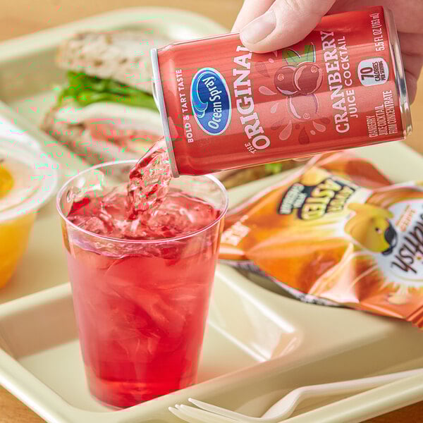A hand pouring Ocean Spray Cranberry Juice from a red can into a glass of red liquid.