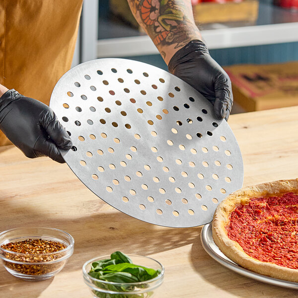 A round, perforated metal pizza disk being held above a pizza and some ingredients on a wooden counter.