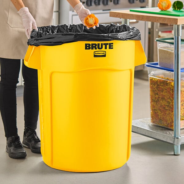 A woman standing next to a yellow Rubbermaid BRUTE trash can.