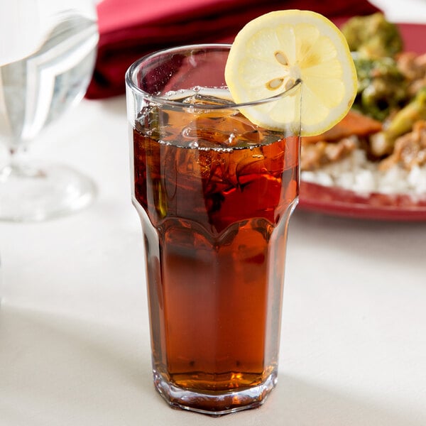 A clear Carlisle plastic tumbler filled with brown liquid and ice with a lemon slice on top.