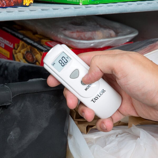 A hand using a Taylor digital infrared thermometer to check a refrigerator temperature.