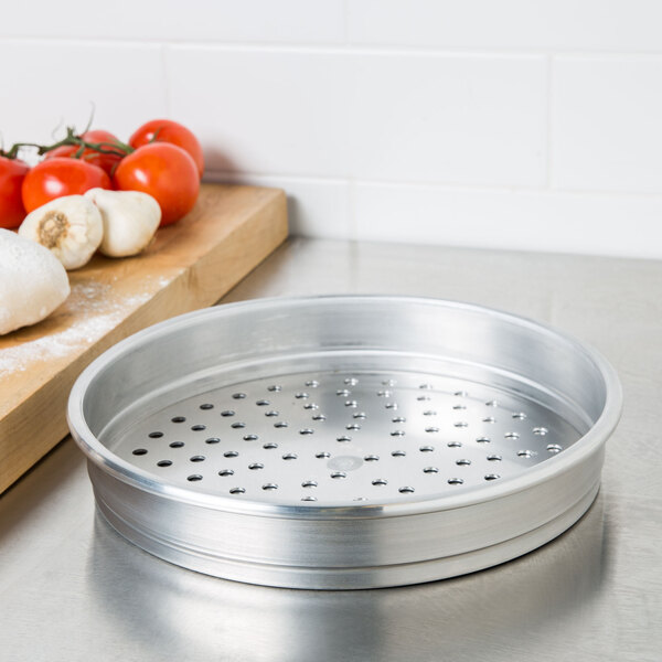 An American Metalcraft heavy weight aluminum pizza pan with holes on the bottom sitting on a table next to tomatoes and garlic.