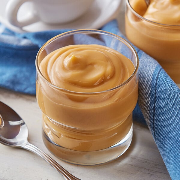 A glass of Cafe Classics Butterscotch Pudding with a spoon on a table with a blue napkin.