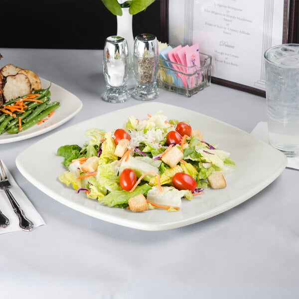 A Tuxton eggshell white china plate with salad on it on a table.