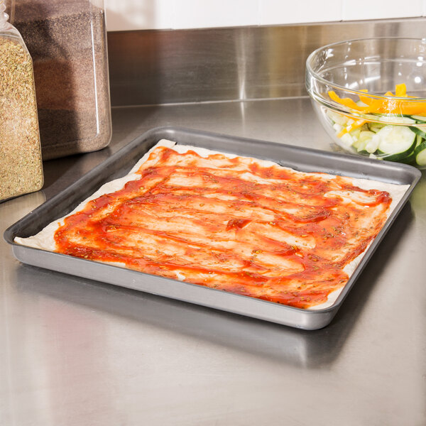 A pizza with sauce on an American Metalcraft square pizza pan on a counter.