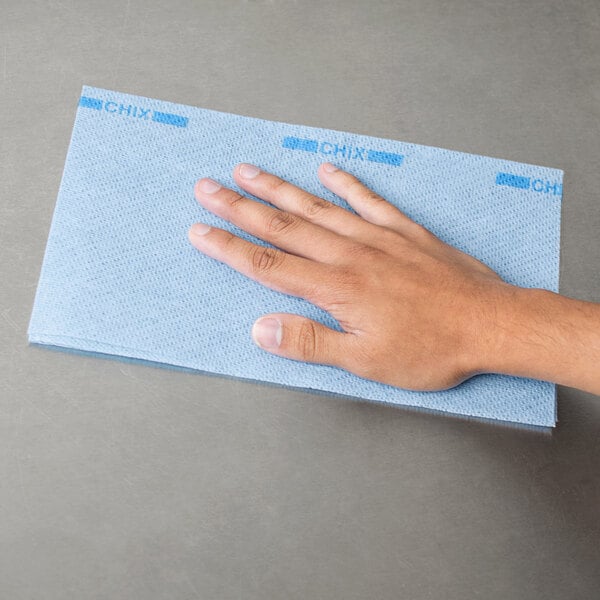 A person's hand using a blue Chicopee foodservice towel on a table.