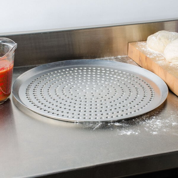 An American Metalcraft heavy weight aluminum pizza pan with dough on it next to a cutting board and a container of liquid.