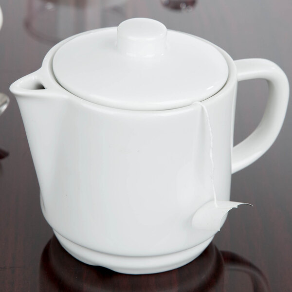An Arcoroc white porcelain teapot on a table with a tea bag attached.
