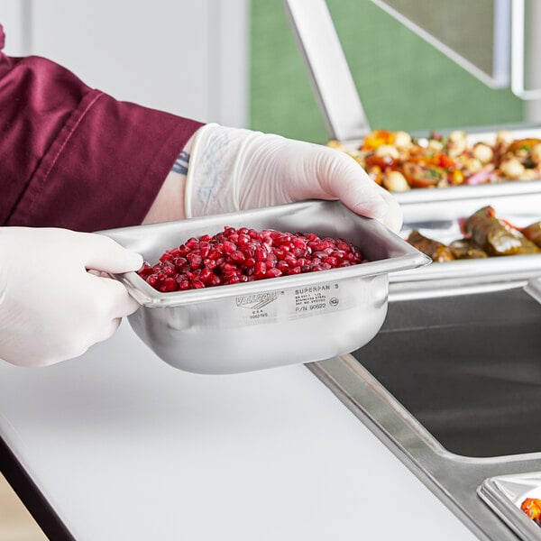 A person in gloves holding a Vollrath stainless steel steam table pan full of food.