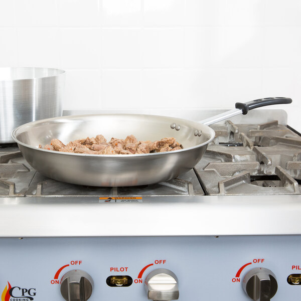 A Vollrath stainless steel fry pan with black silicone handle cooking on a stove.