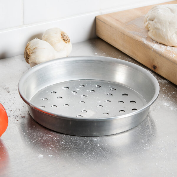 An American Metalcraft tin-plated steel pizza pan with holes next to garlic on a cutting board.