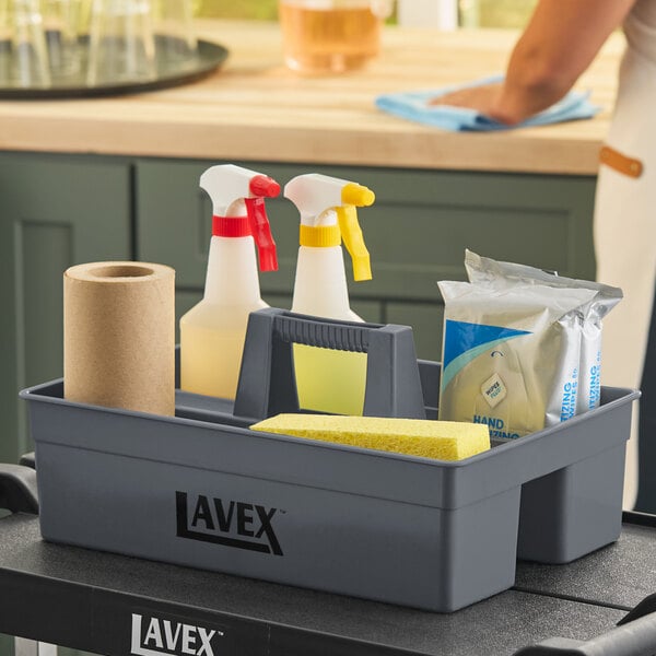 A gray plastic 3-compartment cleaning caddy containing cleaning supplies such as spray bottles, a sponge, a roll of paper towels, and sanitizing wipes.