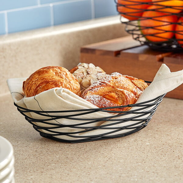 A Tablecraft black wire basket filled with croissants and pastries on a counter.